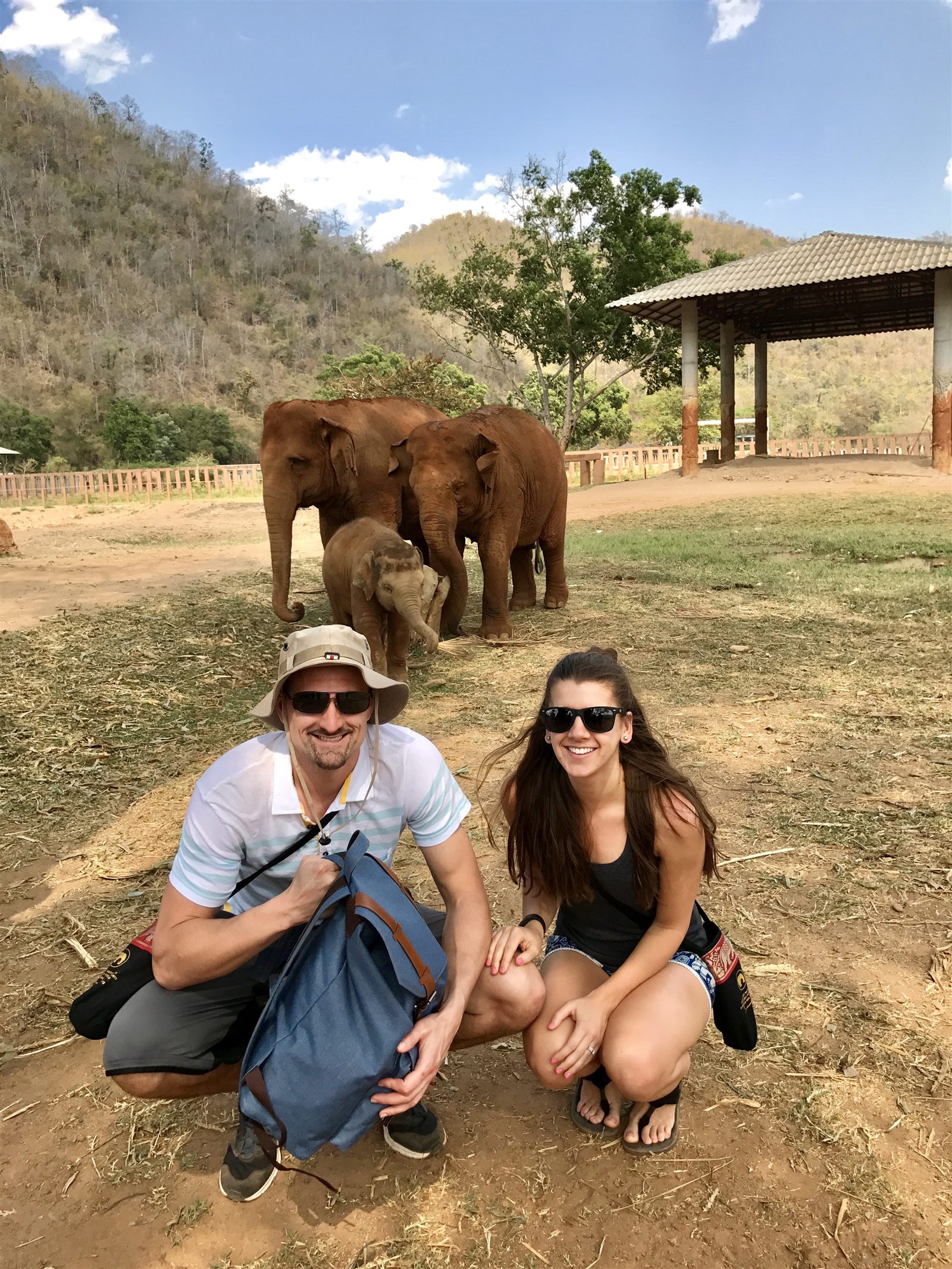 Kyle and Kailah crouching near a family of elephants at Elephant Nature Park in Chiang Mai